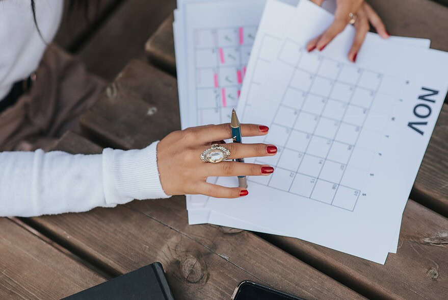 Calendar for November on desk as a woman flips the page up slightly to add items to the calendar. You can only see the woman's hand and fore arm.